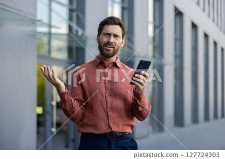 A man in a red shirt is holding a cell phone. He is looking at the phone and he is confused 127724303