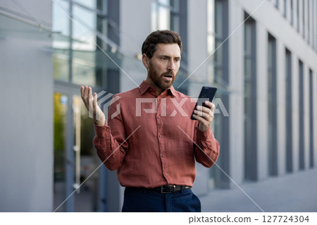 Mature male professional standing outside in city street holding phone, expression of shock and disbelief. Businessman in smart casual attire reacting to surprising news Mature male professional standing outside in city street holding phone, expression of shock and disbelief. Businessman in smart casual attire reacting to surprising news 127724304