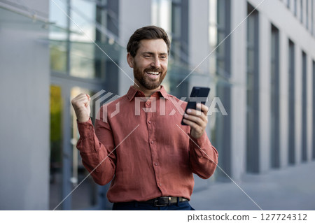 Mature man outdoors expressing excitement after obtaining good news on phone. He celebrates success outside modern office building in city, holding smartphone, smiling with positive energy. 127724312
