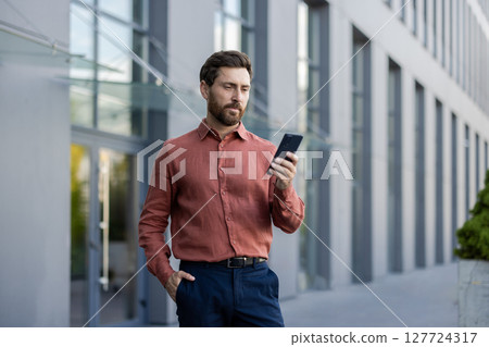 A man in a red shirt is looking at his cell phone. He is standing in front of a building A man in a red shirt is looking at his cell phone. He is standing in front of a building 127724317
