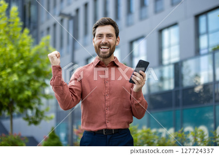 Mature man celebrating winning moment holding smartphone outdoors. Expressing excitement, success, and happiness with confident smile in urban background. Concept of joy and achievement with phone. Mature man celebrating winning moment holding smartphone outdoors. Expressing excitement, success, and happiness with confident smile in urban background. Concept of joy and achievement with phone. 127724337