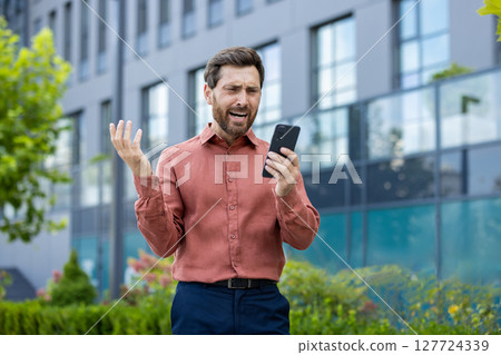 Frustrated mature businessman outdoors holding phone in hand, expressing exasperation. Wearing casual pink shirt, raising arms in dissatisfaction, standing near modern glass office building. Frustrated mature businessman outdoors holding phone in hand, expressing exasperation. Wearing casual pink shirt, raising arms in dissatisfaction, standing near modern glass office building. 127724339