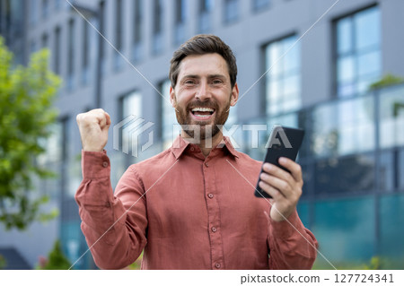 Mature man expressing joy holding smartphone outdoors near modern building. Celebrating success, positive news. Casual style, business victory, urban environment, cheerful and thrilled. Mature man expressing joy holding smartphone outdoors near modern building. Celebrating success, positive news. Casual style, business victory, urban environment, cheerful and thrilled. 127724341