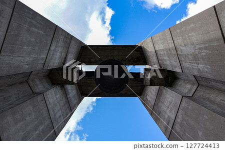 A large dark bell hangs prominently silhouetted within a monumental brutalist concrete structure, viewed from below against a vibrant blue sky, conveying remembrance and history A large dark bell hangs prominently silhouetted within a monumental brutalist concrete structure, viewed from below against a vibrant blue sky, conveying remembrance and history 127724413