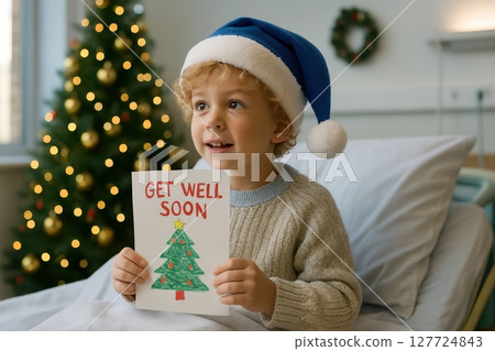 Smiling boy in blue santa hat holding get well soon card in hospital room with christmas tree 127724843