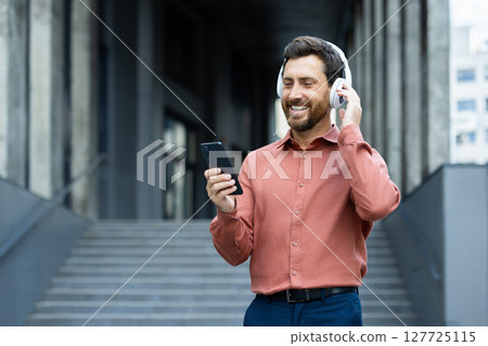 A smiling man adjusts his headphones while looking at his smartphone, enjoying music. He wears a casual shirt in an urban environment. A smiling man adjusts his headphones while looking at his smartphone, enjoying music. He wears a casual shirt in an urban environment. 127725115