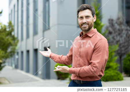 A smiling man in shirt stands in front of a modern building with outstretched hands, inviting viewers in 127725185