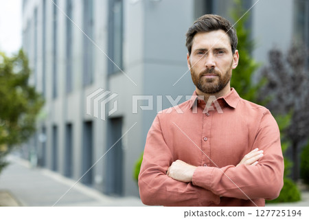 A confident man with a beard stands with arms crossed, looking directly at the camera in an urban setting. 127725194