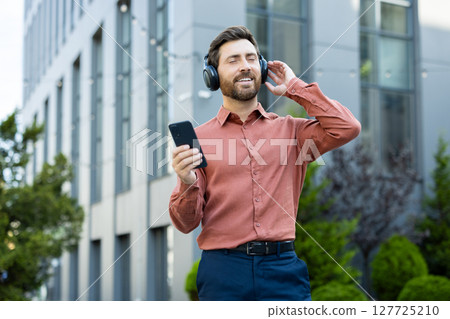 A man wearing headphones listens to music on his phone outside a modern building, enjoying the moment with his eyes closed. 127725210
