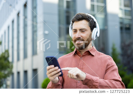 A man with headphones uses his phone outside on a city street with modern buildings, enjoying music or connecting with others A man with headphones uses his phone outside on a city street with modern buildings, enjoying music or connecting with others 127725223