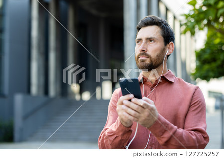 A man in a stylish shirt enjoys music from his smartphone while standing outside in the bright sunlight. 127725257
