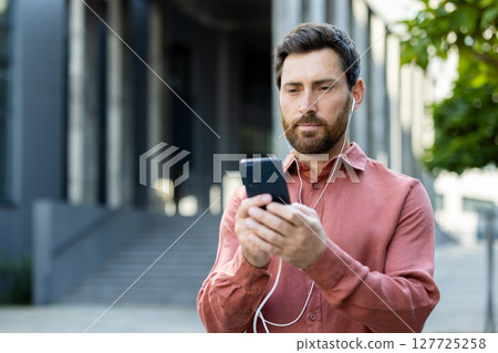 A man with a beard stands outside a modern building listening to music, looking at his mobile phone in hand on a sunny day. 127725258