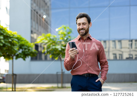 A well-dressed man listens to music or communicates on his phone while outdoors. The sunlight catches his smiling face as he enjoys a moment. A well-dressed man listens to music or communicates on his phone while outdoors. The sunlight catches his smiling face as he enjoys a moment. 127725261