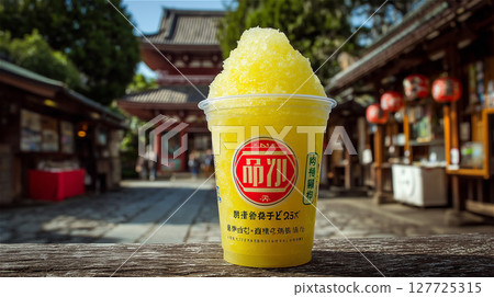 Shaved ice with yellow syrup served in a clear cup with a traditional shrine grounds in the background 127725315
