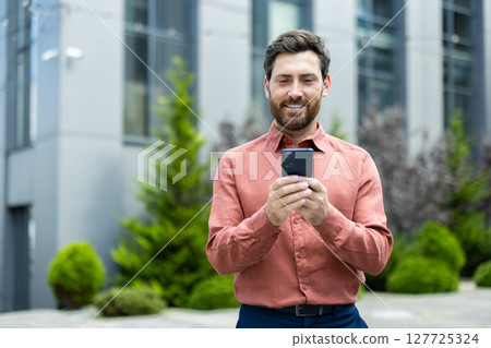 Successful man walking through the city outside an office building with a phone in his hands. Businessman using an app on his smartphone, typing messages, and browsing social media, smiling. Successful man walking through the city outside an office building with a phone in his hands. Businessman using an app on his smartphone, typing messages, and browsing social media, smiling. 127725324