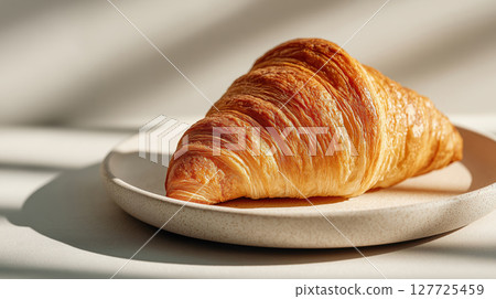 Golden croissant with flaky layers served on a ceramic plate in soft natural light. Buttery and crisp pastry with delicate texture. Morning breakfast scene. 127725459