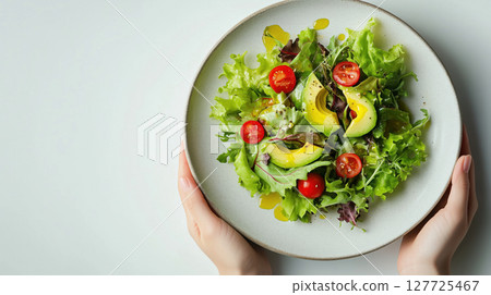 Fresh vegetable salad with avocado slices, cherry tomatoes, and mixed greens served on a ceramic plate held by hands. Healthy and colorful food. Clean eating. 127725467