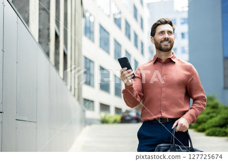 A well-dressed man walks outside, holding a phone in one hand and a briefcase in the other, smiling. 127725674