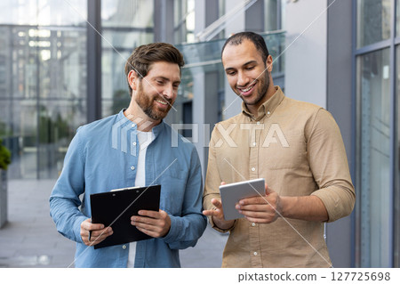 Two diverse businessmen review a tablet, possibly discussing a project or presenting ideas, smiling in a modern outdoor setting. Two diverse businessmen review a tablet, possibly discussing a project or presenting ideas, smiling in a modern outdoor setting. 127725698