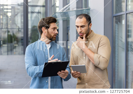 Two men are engaged in a discussion outdoors. One holds a clipboard, while the other considers a tablet in their hands. Two men are engaged in a discussion outdoors. One holds a clipboard, while the other considers a tablet in their hands. 127725704