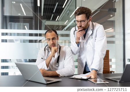 Two doctors, wearing white coats and stethoscopes, look at a laptop screen in a modern office setting, deep in thought. Two doctors, wearing white coats and stethoscopes, look at a laptop screen in a modern office setting, deep in thought. 127725711