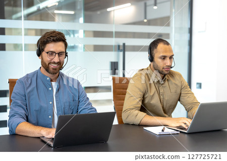 Two customer service representatives wearing headsets are working at desks using their laptops in a modern office setting. 127725721