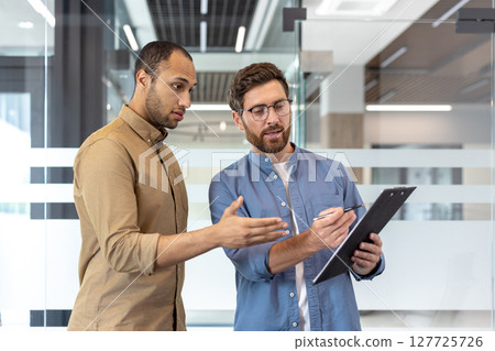 Two business professionals discuss paperwork, one pointing and the other holding a pen, in a modern office setting. 127725726