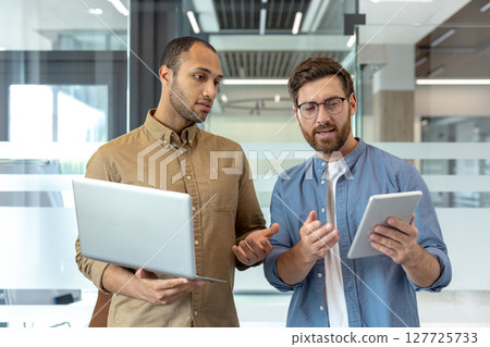 Two professional men collaborate in a modern office, using a laptop and tablet for discussion. They appear focused and engaged in their work. Two professional men collaborate in a modern office, using a laptop and tablet for discussion. They appear focused and engaged in their work. 127725733
