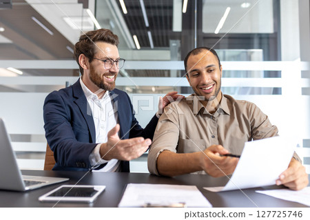 Two diverse businessmen smiling, reviewing paperwork, and discussing matters in a modern office environment. Collaboration and teamwork are evident. Two diverse businessmen smiling, reviewing paperwork, and discussing matters in a modern office environment. Collaboration and teamwork are evident. 127725746