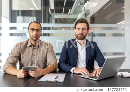 Two confident business professionals sit at a desk, appearing to be ready to work together in a modern office. One uses a laptop. Two confident business professionals sit at a desk, appearing to be ready to work together in a modern office. One uses a laptop. 127725751