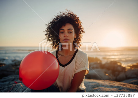 A woman with curly hair is sitting on a rock with a red balloon in her hand 127725887