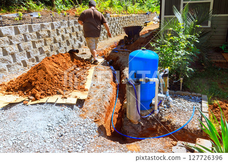 Homeowner works on installing water well pump system near trench in thoughtfully arranged outdoor space. 127726396