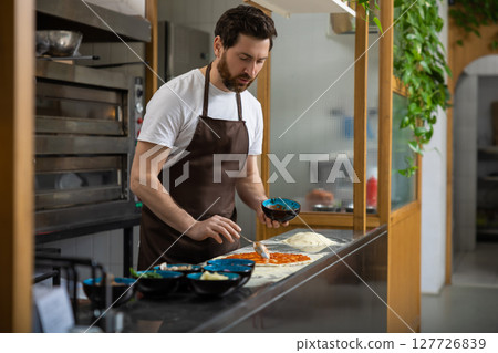 Male cooker working in the pizzeria kitchen and putting topping to the pizza Male cooker working in the pizzeria kitchen and putting topping to the pizza 127726839