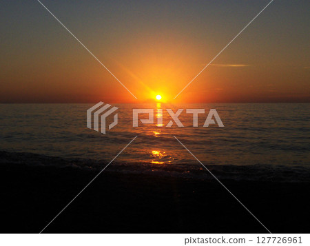 The breathtaking beauty of a sunset in Calabria, Italy. The sky is painted in rich hues of orange, pink, and gold as the sun dips below the horizon, casting a warm glow over the calm sea. 127726961