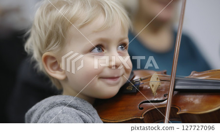 A young boy with blonde hair smiles while holding a violin, engaging in a music class at a community center. His concentration highlights the joy of learning an instrument. 127727096