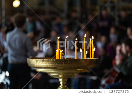 Burning candles on a golden plate during a ceremony with blurred people in the background 127727275