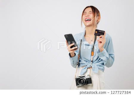 Travel vibe with travel-ready Traveler tourist Asian woman using app on phone. She excited smiles holding photo camera and credit card bank while booking her vacation online. isolated white background 127727384