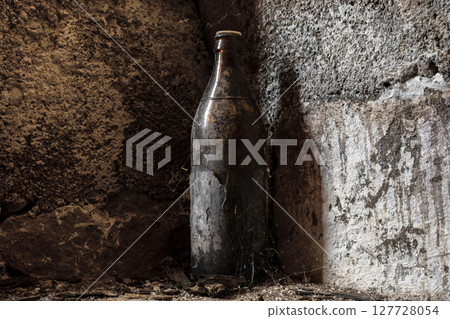 Very old and aged brown bottle covered with dust and spider web standing in front of the concrete wall at basement 127728054