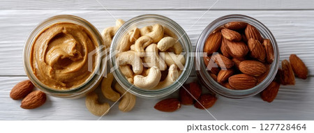 Different types of peanut butter are surrounded by nuts on a light wooden table, highlighted by soft, sunny light and pastel hues 127728464