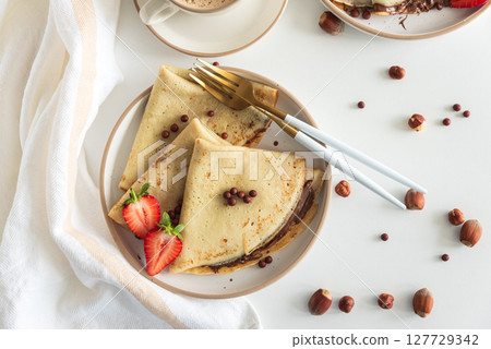 Crepes with chocolate hazelnut spread, strawberries and hazelnuts on a white wooden table. Top view 127729342