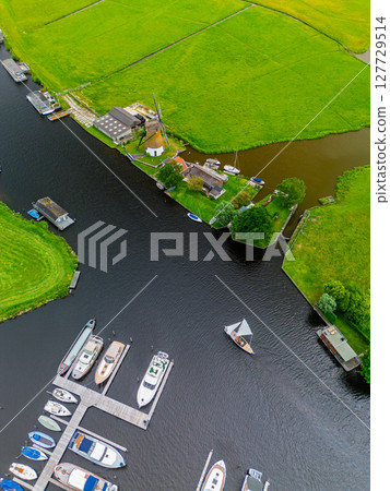 A top-down view of a marina with multiple boats docked along wooden piers, a boat cruising through a narrow waterway surrounded by green parks and natural grassy areas. A top-down view of a marina with multiple boats docked along wooden piers, a boat cruising through a narrow waterway surrounded by green parks and natural grassy areas. 127729514