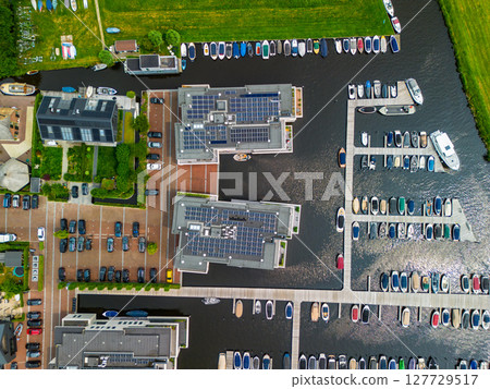 A top-down view of a marina with multiple boats docked along wooden piers, a boat cruising through a narrow waterway surrounded by green parks and natural grassy areas. Solar panels. 127729517
