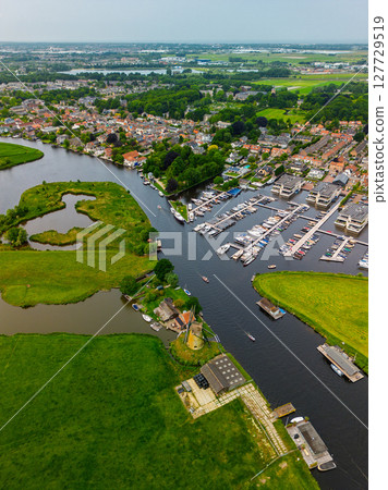 A top-down view of a marina with multiple boats docked along wooden piers, a boat cruising through a narrow waterway surrounded by green parks and natural grassy areas. 127729519