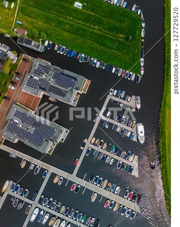 A top-down view of a marina with multiple boats docked along wooden piers, a boat cruising through a narrow waterway surrounded by green parks and natural grassy areas. Solar panels. A top-down view of a marina with multiple boats docked along wooden piers, a boat cruising through a narrow waterway surrounded by green parks and natural grassy areas. Solar panels. 127729520