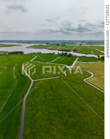 Aerial view of a wide green landscape with winding canals dividing the fields. Cows graze on the pastures, and a large river flows in the distance under a cloudy sky. Peaceful rural scenery. 127729521