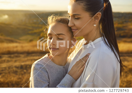 Young mother embracing her teenage child girl outdoor standing with closed eyes in the field. 127729743