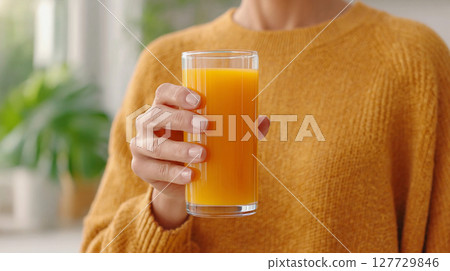Freshly squeezed orange juice in a tall glass held by a person wearing a cozy sweater in a bright indoor setting with greenery in the background 127729846