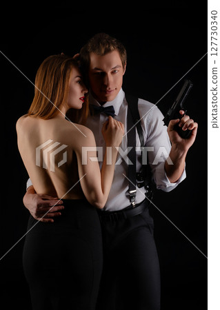 Couple in elegant attire poses dramatically with a gun in a dark setting Couple in elegant attire poses dramatically with a gun in a dark setting 127730340