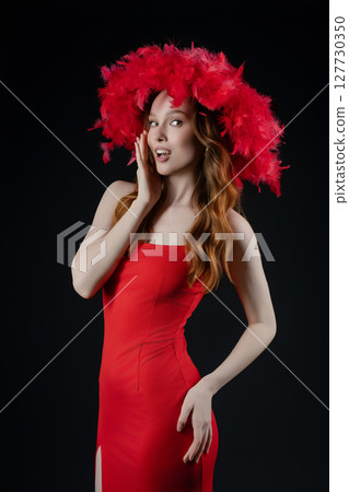 Confident woman poses in studio wearing red dress and dramatic feathered headdress, expressing playful emotion in artistic shoot 127730350