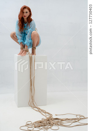 Red-haired woman poses elegantly in rope bondage on a white backdrop 127730403